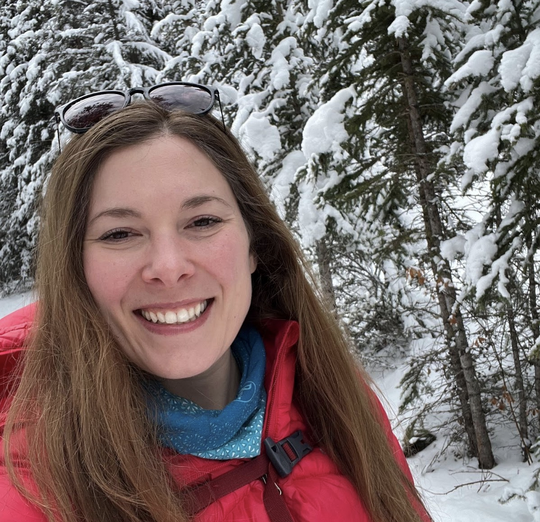 A picture of Laura Kroeker in a red winter jacket smiling in front of a forest covered in snow.